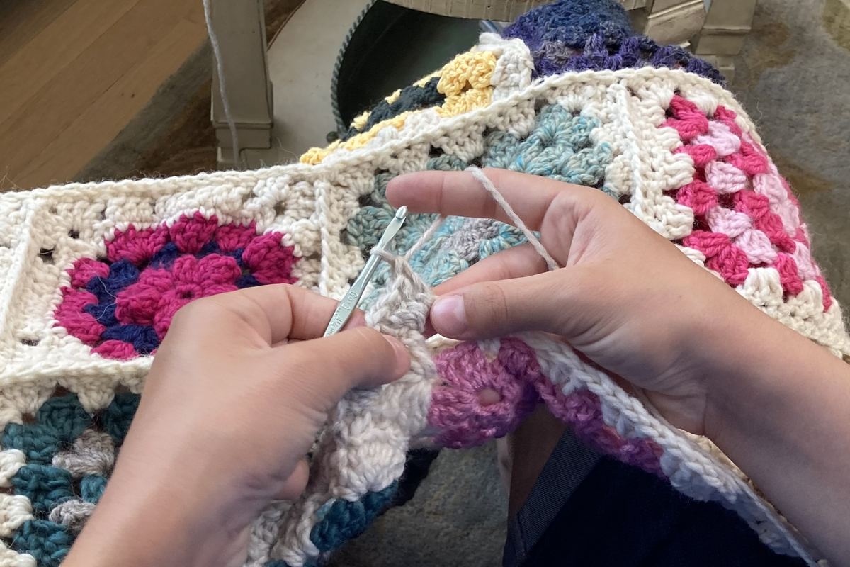 Dos manos tejiendo a crochet con una aguja fina sobre una labor de cuadros granny a medio hacer, en rosa, azul y crudo