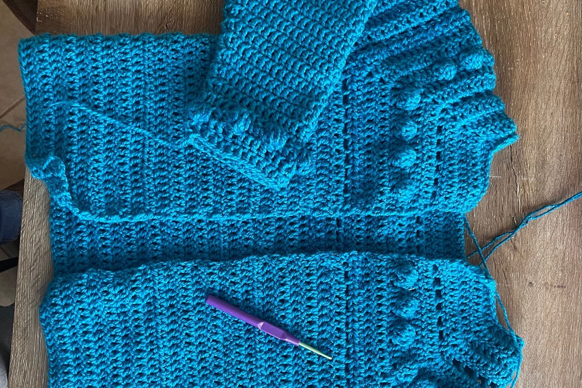 Un cárdigan pequeño de crochet en azul petróleo, extendido en plano sobre una mesa de madera con una manga doblada y la orilla de bolitas del puño a la vista. Encima hay un crochet morado, que da la escala